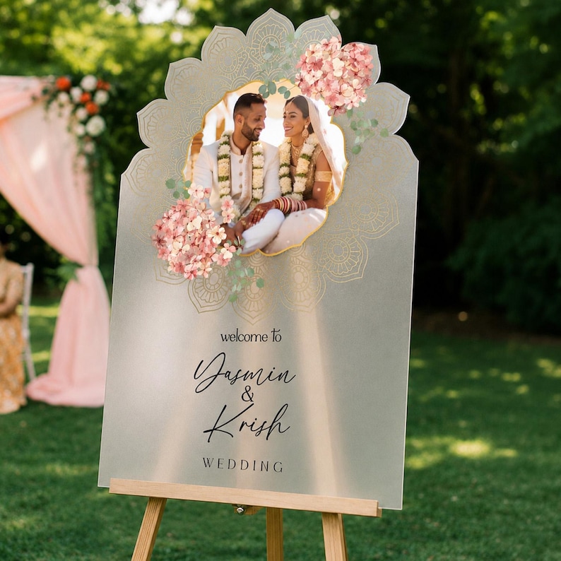 Peut inclure: Un panneau de bienvenue de mariage en acrylique d&eacute;poli avec une photo centrale d'un couple en tenue traditionnelle, orn&eacute; d'illustrations d'hortensias roses et de motifs mandala dor&eacute;s. Le texte indique, welcome to Yasmin & Krish WEDDING.