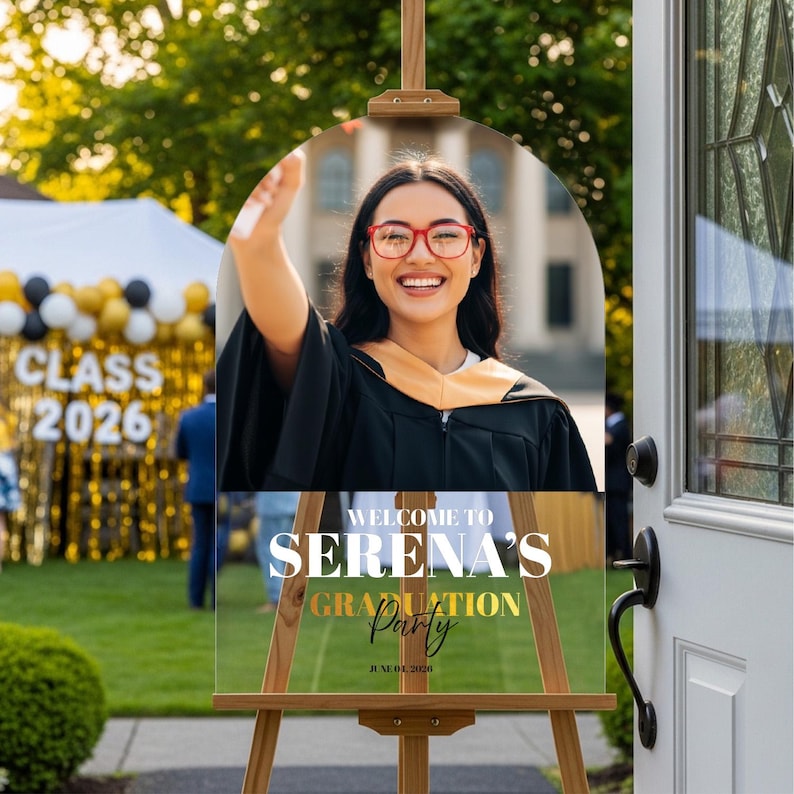 May include: A clear acrylic graduation party sign on a wooden easel. The sign features a photo of a smiling graduate wearing a cap and gown. Text on the sign reads "Welcome to Serena's Graduation Party" and "June 01, 2026".