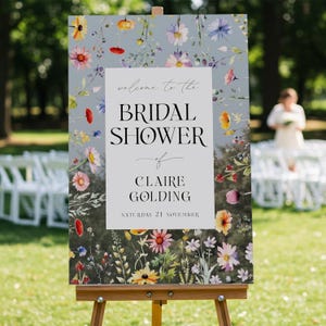 May include: A rectangular sign on a wooden easel featuring a white center panel with the text "welcome to the BRIDAL SHOWER of CLAIRE GOLDING SATURDAY 21 NOVEMBER" surrounded by a border of colorful watercolor wildflowers on a gray background.