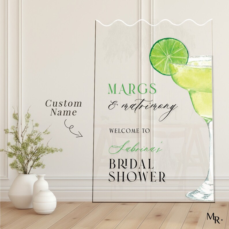 May include: A clear acrylic sign with a green and white margarita illustration. The text on the sign reads "Margs & Matrimony, Welcome to Sabrina's Bridal Shower".
