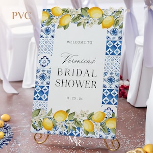 May include: A rectangular PVC welcome sign for a bridal shower featuring a white centre with black text reading WELCOME TO Veronica's BRIDAL SHOWER 11.25.24. The border displays blue and white tile patterns, yellow lemons, and white blossoms.