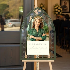 May include: A memorial sign on a wooden easel. The sign features a photo of a woman in a green top, framed by a decorative green arch. Text on the sign reads "IN THE MEMORY OF Amelia Ford 1975 - 2023". The background shows a church interior.