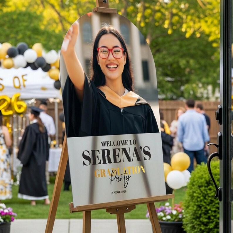 May include: A graduation party sign on a wooden easel. The sign features a photo of a smiling woman in a graduation gown and glasses. The sign reads "Welcome to Serena's Graduation Party" with the date June 01, 2026.