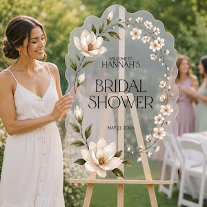 May include: A scalloped, transparent acrylic sign on a wooden easel featuring a floral wreath of white magnolia and cherry blossoms. The text reads: WELCOME TO HANNAH'S BRIDAL SHOWER MAY 21, 2026. The design includes green leaves and stems.