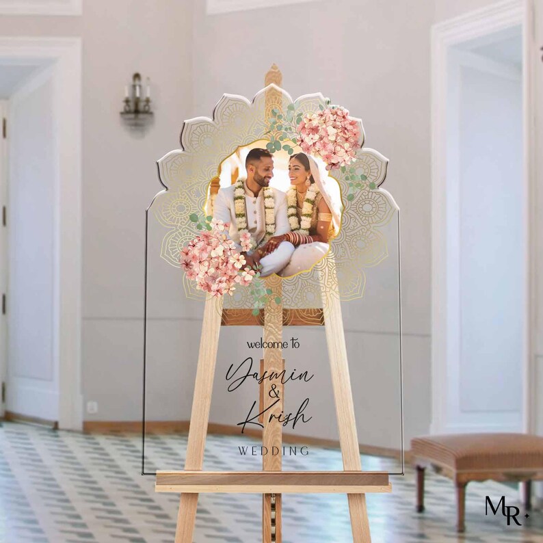 Peut inclure: Un panneau de bienvenue de mariage en acrylique transparent avec un bord festonn&eacute;, pr&eacute;sentant une photo centrale d'un couple en tenue traditionnelle, orn&eacute; d'illustrations florales roses et de motifs mandala dor&eacute;s, avec le texte "welcome to Yasmin & Krish WEDDING".