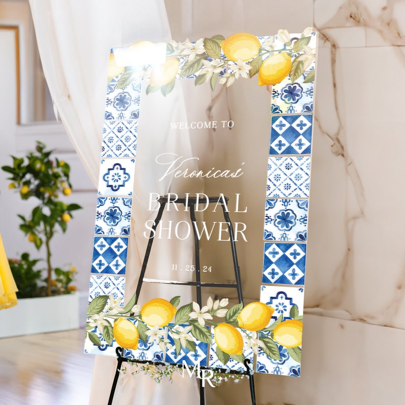 May include: A rectangular clear acrylic sign featuring a border of blue and white Mediterranean-style tiles, yellow lemons, and white blossoms. The text reads: WELCOME TO Veronica's BRIDAL SHOWER 11.25.24. The sign is displayed on a black metal easel.