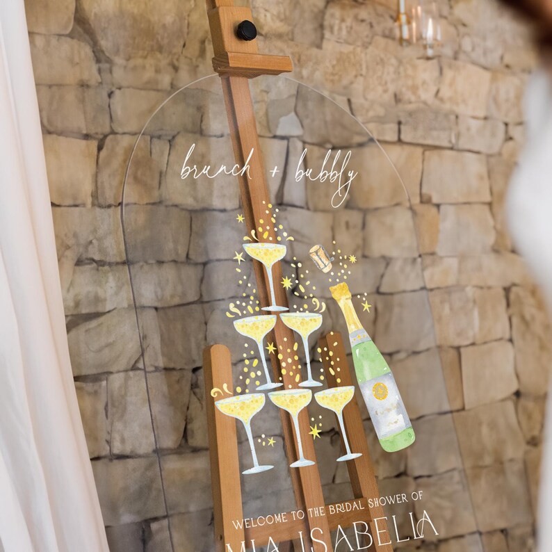 May include: A clear acrylic sign with a champagne bottle and glasses illustration. The sign reads "brunch + bubbly" and "Welcome to the bridal shower of Mia Isabella".