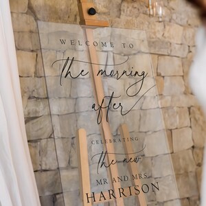 K&ouml;nnte beinhalten: Klares Acrylschild mit schwarzem Kalligraphietext, der "Willkommen zum Morgen danach, um das neue Herr und Frau Harrison zu feiern" lautet.