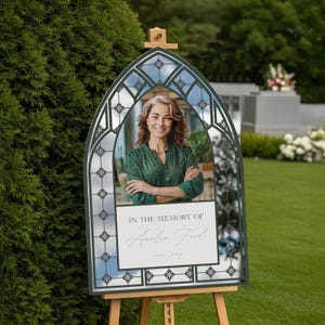 May include: A memorial sign with a photo of a woman, framed by a dark green, arched structure with a stained-glass design. The sign reads "In the Memory of Amelia Ford" with the years 1973-2023. The sign is displayed on a wooden easel outdoors.