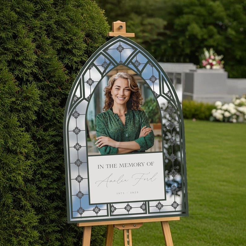 May include: A memorial sign with a photo of a woman, framed by a dark green, arched structure with a stained-glass design. The sign reads "In the Memory of Amelia Ford" with the years 1973-2023. The sign is displayed on a wooden easel outdoors.