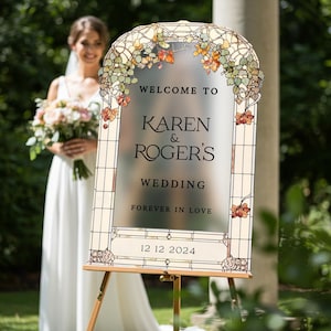 May include: An arched wedding welcome sign with a frosted center and stained glass border featuring autumn leaves. The text reads: WELCOME TO KAREN & ROGER'S WEDDING FOREVER IN LOVE 12 12 2024. The sign is mounted on a wooden easel stand.