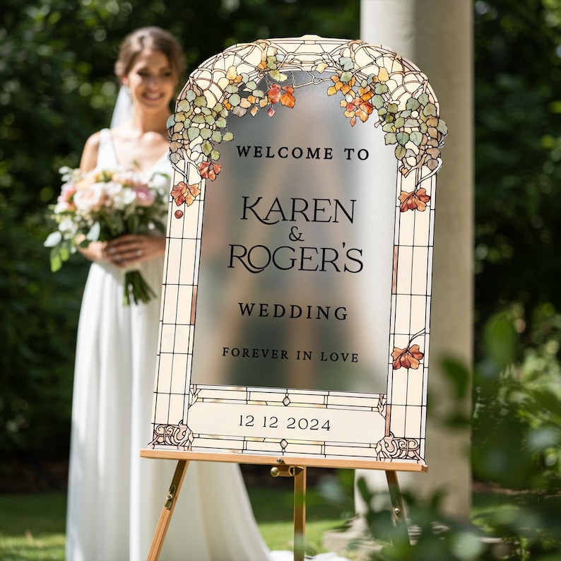 May include: An arched wedding welcome sign with a frosted center and stained glass border featuring autumn leaves. The text reads: WELCOME TO KAREN & ROGER'S WEDDING FOREVER IN LOVE 12 12 2024. The sign is mounted on a wooden easel stand.