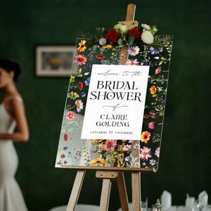 May include: A clear acrylic sign on a wooden easel featuring a colorful floral border with red, yellow, and purple wildflowers. The center text reads: welcome to the BRIDAL SHOWER of CLAIRE GOLDING SATURDAY 21 NOVEMBER. A floral arrangement sits on top.