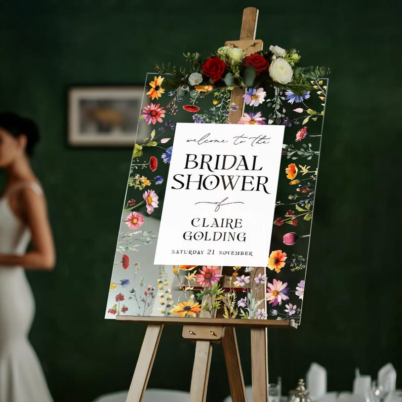 May include: A clear acrylic sign on a wooden easel featuring a colorful floral border with red, yellow, and purple wildflowers. The center text reads: welcome to the BRIDAL SHOWER of CLAIRE GOLDING SATURDAY 21 NOVEMBER. A floral arrangement sits on top.