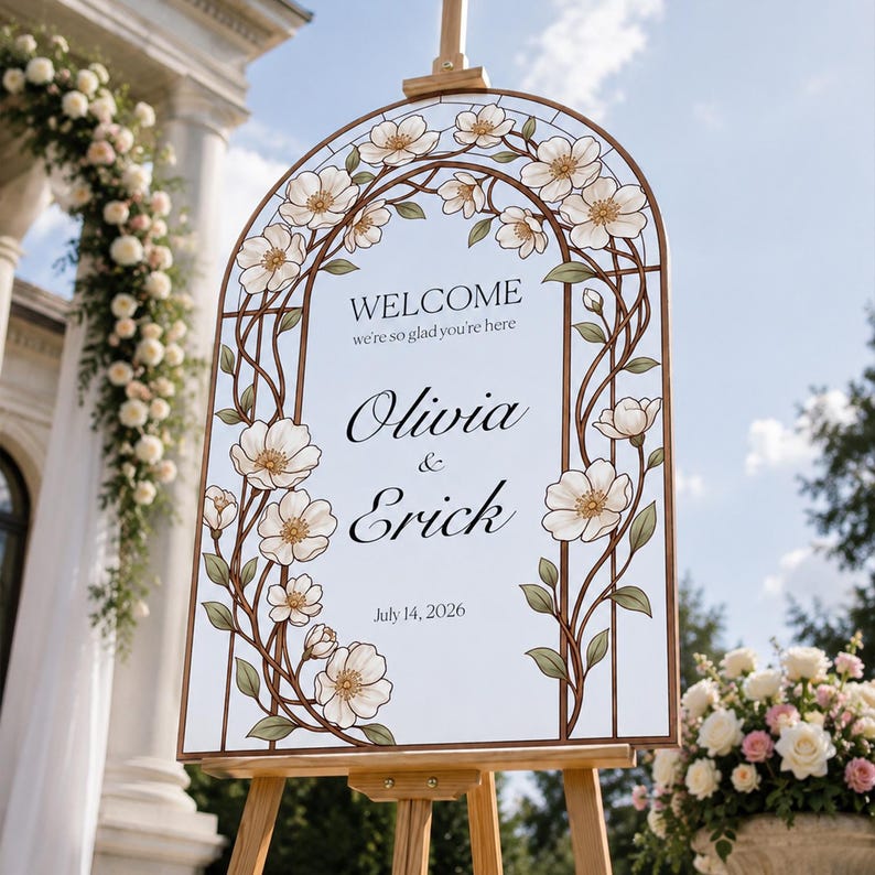 K&ouml;nnte beinhalten: Ein bogenf&ouml;rmiges Hochzeits-Willkommensschild auf einer Holzstaffelei mit wei&szlig;em Hintergrund und schwarzem Text, der Welcome we're so glad you're here, Olivia & Erick, und July 14, 2026 lautet, umrahmt von einer dekorativen Bord&uuml;re aus wei&szlig;en Blumen.