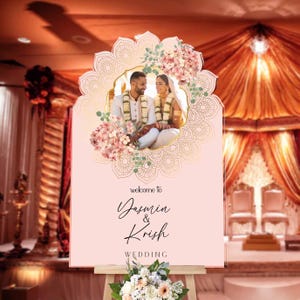 Peut inclure: Un panneau de bienvenue de mariage rectangulaire rose clair avec une photo centrale d'un couple en tenue traditionnelle, encadr&eacute; par un motif mandala dor&eacute; et des fleurs roses. Le texte indique : welcome to Yasmin & Krish WEDDING.