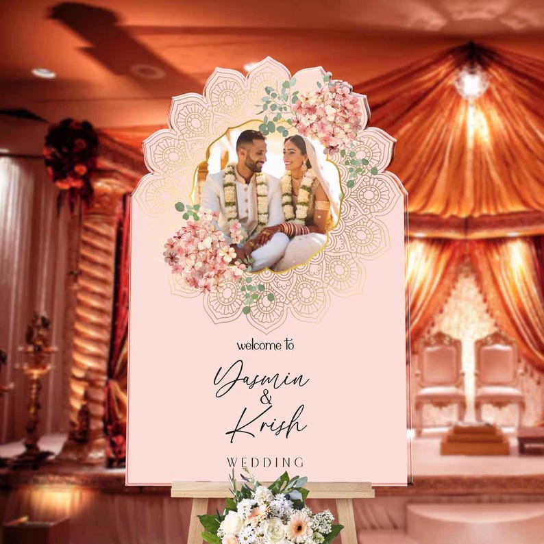 Peut inclure: Un panneau de bienvenue de mariage rectangulaire rose clair avec une photo centrale d'un couple en tenue traditionnelle, encadr&eacute; par un motif mandala dor&eacute; et des fleurs roses. Le texte indique : welcome to Yasmin & Krish WEDDING.