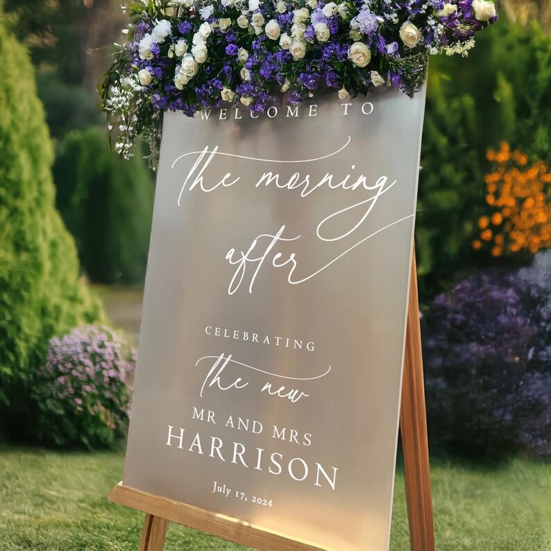 K&ouml;nnte beinhalten: Ein wei&szlig;es Schild mit silbernem Hintergrund auf einem Staffelei mit lila Blumen. Das Schild lautet "Willkommen zum Morgen danach, um das neue Ehepaar Harrison zu feiern, 17. Juli 2024".
