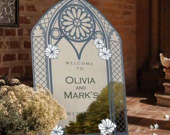 May include: An arched wedding welcome sign featuring a blue gothic window frame design with a rose window motif at the top. The sign displays the text Welcome to Olivia and Mark's Wedding and is decorated with white floral illustrations on the edges.
