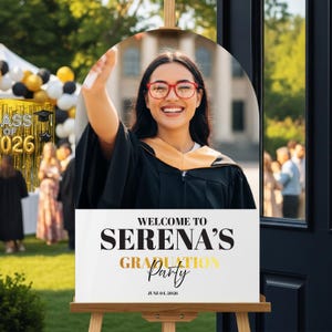 May include: A graduation party welcome sign on an easel. The sign features a photo of a smiling graduate in a cap and gown. Text on the sign reads "Welcome to Serena's Graduation Party, June 01, 2026."