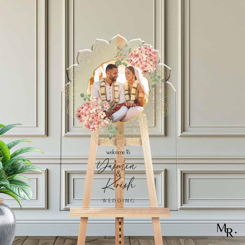 Peut inclure: Un panneau de bienvenue de mariage en acrylique transparent avec un bord sup&eacute;rieur festonn&eacute;, des motifs de mandala dor&eacute;s, des grappes d'hortensias roses et une photo centrale d'un couple en tenue traditionnelle. Texte : welcome to Jasmin & Krish WEDDING.