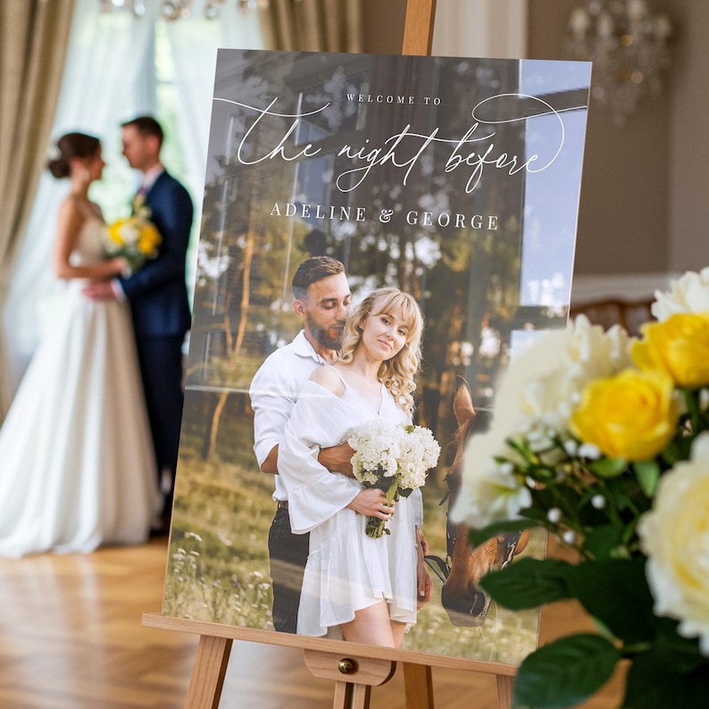 May include: A rectangular wedding welcome sign on a wooden easel featuring a photograph of a couple with a horse. The text reads: Welcome to The night before Adeline & George. The sign is displayed in a room with a blurred couple in the background.