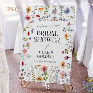 May include: A rectangular PVC bridal shower sign featuring a white background with a colorful watercolor wildflower border. The text reads: welcome to the BRIDAL SHOWER of CLAIRE GOLDING SATURDAY 21 NOVEMBER. The sign rests on a gold metal stand.