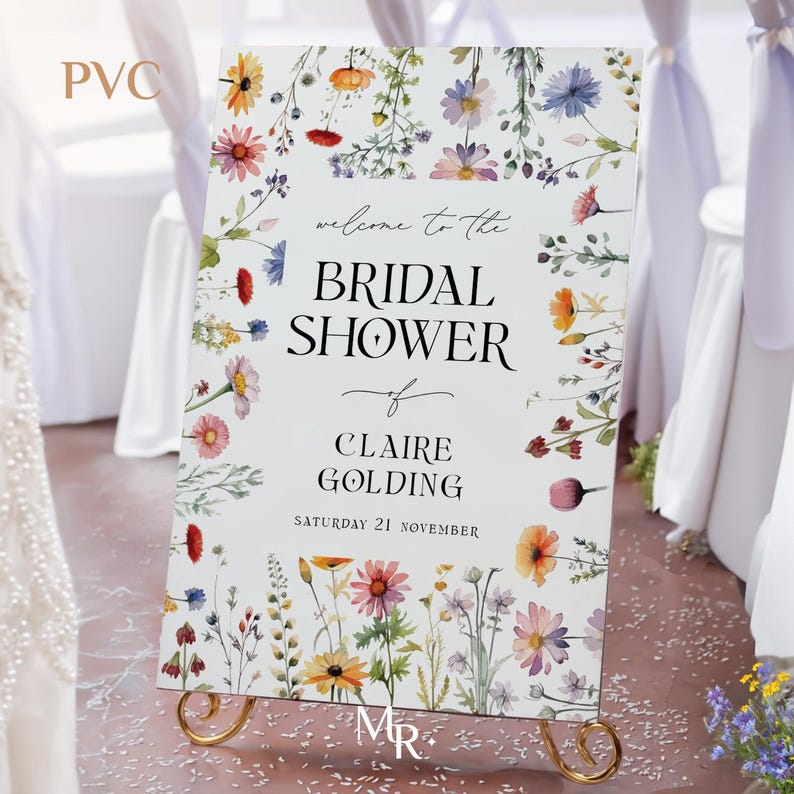 May include: A rectangular PVC bridal shower sign featuring a white background with a colorful watercolor wildflower border. The text reads: welcome to the BRIDAL SHOWER of CLAIRE GOLDING SATURDAY 21 NOVEMBER. The sign rests on a gold metal stand.