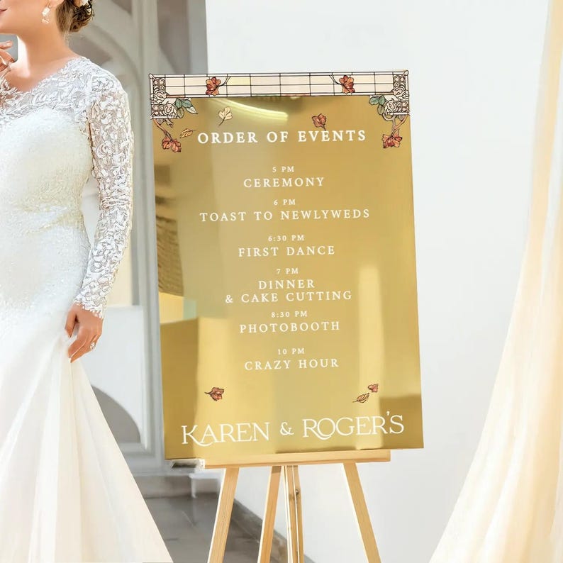 May include: Gold mirror sign with a white frame and black text listing the order of events for a wedding. The sign reads "ORDER OF EVENTS" and lists the times for the ceremony, toast to newlyweds, first dance, dinner and cake cutting, photo booth, and crazy hour. The sign is on a wooden easel and has the names "KAREN & ROGER'S" at the bottom.