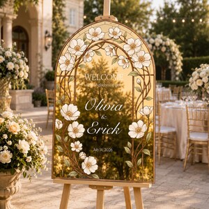 K&ouml;nnte beinhalten: Ein bogenf&ouml;rmiges Hochzeits-Willkommensschild mit einem Rand im Buntglas-Stil aus wei&szlig;en Bl&uuml;ten und gr&uuml;nen Ranken. Die goldfarbene Oberfl&auml;che zeigt den Text: WELCOME we're so glad you're here, Olivia & Erick, sowie das Datum 14. Juli 2026.