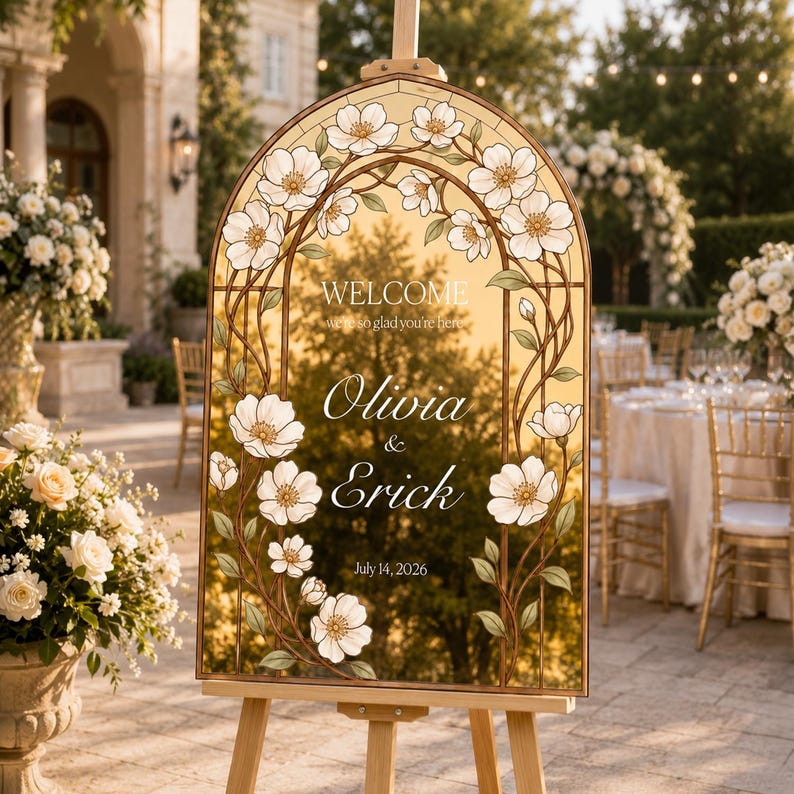 K&ouml;nnte beinhalten: Ein bogenf&ouml;rmiges Hochzeits-Willkommensschild mit einem Rand im Buntglas-Stil aus wei&szlig;en Bl&uuml;ten und gr&uuml;nen Ranken. Die goldfarbene Oberfl&auml;che zeigt den Text: WELCOME we're so glad you're here, Olivia & Erick, sowie das Datum 14. Juli 2026.