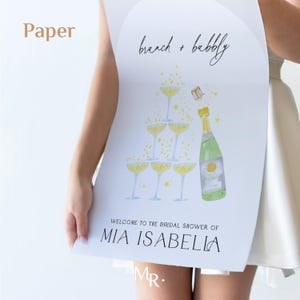 May include: A white sign with the text "brunch + bubbly" and a watercolor illustration of a champagne bottle and glasses. The sign also includes the text "Welcome to the bridal shower of Mia Isabella".