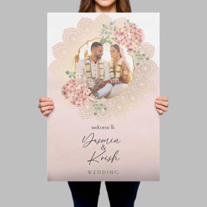 Peut inclure: Un panneau de bienvenue de mariage rectangulaire avec une photo centrale d'un couple en tenue traditionnelle, encadr&eacute;e par un motif mandala dor&eacute; et des fleurs roses. Le texte indique : welcome to Yasmin & Krish WEDDING en noir.