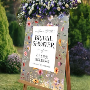 May include: A rectangular sign on a wooden easel featuring a white central panel with the text "welcome to the BRIDAL SHOWER of CLAIRE GOLDING SATURDAY 21 NOVEMBER". The sign has a floral border with colorful wildflowers on a light background.