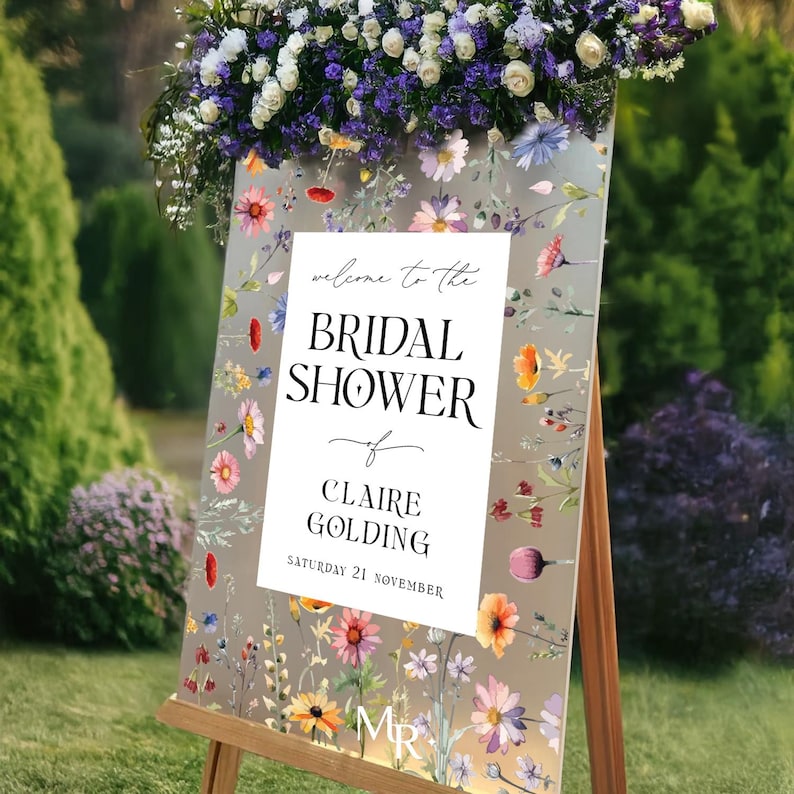 May include: A rectangular sign on a wooden easel featuring a white central panel with the text "welcome to the BRIDAL SHOWER of CLAIRE GOLDING SATURDAY 21 NOVEMBER". The sign has a floral border with colorful wildflowers on a light background.
