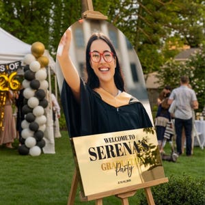 May include: A graduation party welcome sign on a wooden easel. The sign features a photo of a smiling graduate wearing a cap and gown. Gold text reads "Welcome to Serena's Graduation Party" with the date June 01, 2026.