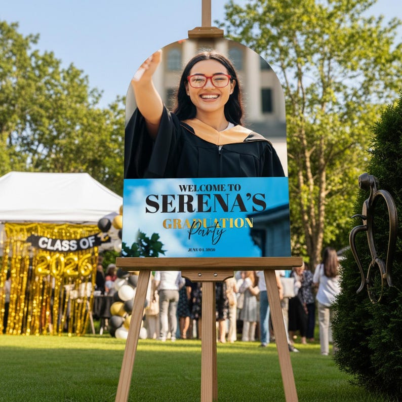 May include: A graduation party sign on a wooden easel. The sign features a photo of a smiling graduate in a cap and gown. Text on the sign reads "Welcome to Serena's Graduation Party" with the date June 04, 2026.