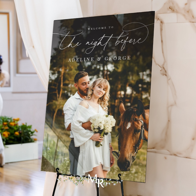 May include: A rectangular wedding welcome sign featuring a photograph of a couple embracing next to a brown horse in a forest. The text reads "WELCOME TO The night before ADELINE & GEORGE" in white script and serif fonts on a dark background.