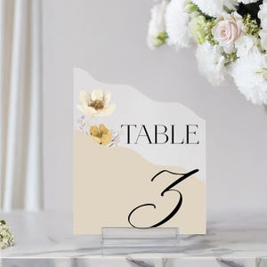 Puede incluir: Una tarjeta de n&uacute;mero de mesa beige con un dise&ntilde;o floral blanco y el texto "TABLE 3" en negro.
