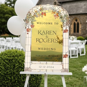 May include: An arched wedding sign with a stained glass border featuring green and orange leaves. The center is gold with the text: WELCOME TO KAREN & ROGER'S WEDDING FOREVER IN LOVE 12 12 2024. The sign is displayed on a white easel stand.