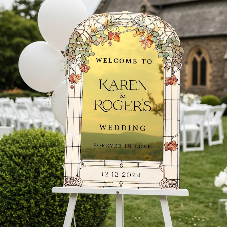 May include: An arched wedding sign with a stained glass border featuring green and orange leaves. The center is gold with the text: WELCOME TO KAREN & ROGER'S WEDDING FOREVER IN LOVE 12 12 2024. The sign is displayed on a white easel stand.