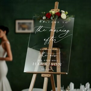 K&ouml;nnte beinhalten: Ein klares Acrylschild mit wei&szlig;er Kalligraphie, auf dem "Willkommen zum Morgen danach, um das neue Ehepaar Harrison zu feiern, 17. Juli 2024" steht. Das Schild steht auf einem Holzstaffelei mit roten und wei&szlig;en Blumen.