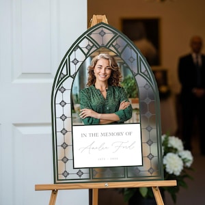 May include: Memorial sign with a photo of a woman in a green blouse, framed by a decorative arch. The sign reads "In the Memory of Amelia Ford 1975-2023" on a white rectangle. The frame is dark green and set on a wooden easel.
