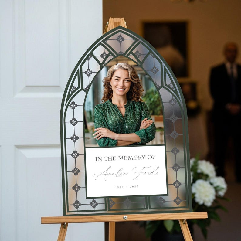 May include: Memorial sign with a photo of a woman in a green blouse, framed by a decorative arch. The sign reads "In the Memory of Amelia Ford 1975-2023" on a white rectangle. The frame is dark green and set on a wooden easel.