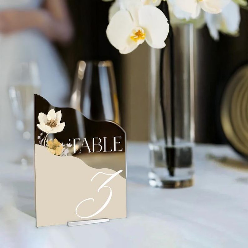 Puede incluir: Un n&uacute;mero de mesa de acr&iacute;lico beige con un dise&ntilde;o floral blanco y la palabra "TABLE" en letras plateadas. El n&uacute;mero "3" est&aacute; escrito en caligraf&iacute;a blanca. El n&uacute;mero de mesa est&aacute; sobre una mesa blanca con un jarr&oacute;n de orqu&iacute;deas blancas en el fondo.