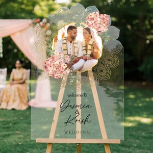 Peut inclure: Un panneau de bienvenue de mariage en acrylique transparent sur un chevalet en bois, orn&eacute; d'une photo centrale d'un couple, de motifs mandala dor&eacute;s et de fleurs roses. Le texte indique : welcome to Yasmin & Krish WEDDING en noir.