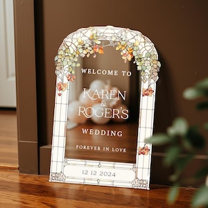 May include: An arched wedding welcome sign with a stained glass border featuring autumn-colored leaves. The text reads: WELCOME TO KAREN & ROGER'S WEDDING FOREVER IN LOVE 12 12 2024. The sign has a clear center panel with a white frame design.
