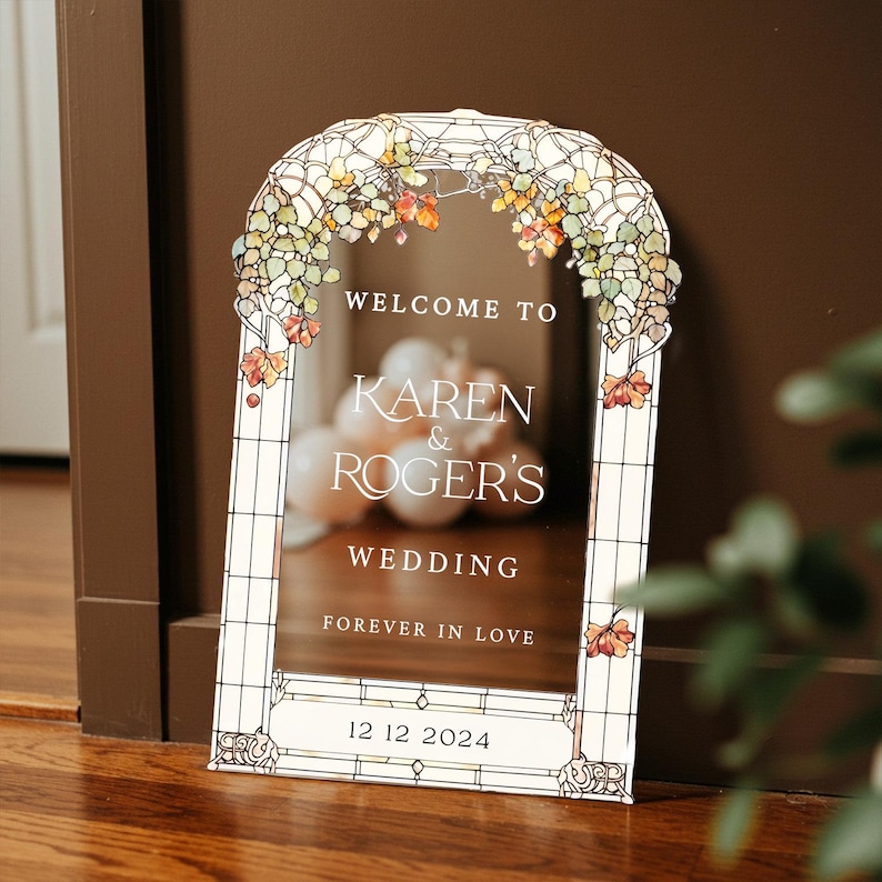 May include: An arched wedding welcome sign with a stained glass border featuring autumn-colored leaves. The text reads: WELCOME TO KAREN & ROGER'S WEDDING FOREVER IN LOVE 12 12 2024. The sign has a clear center panel with a white frame design.