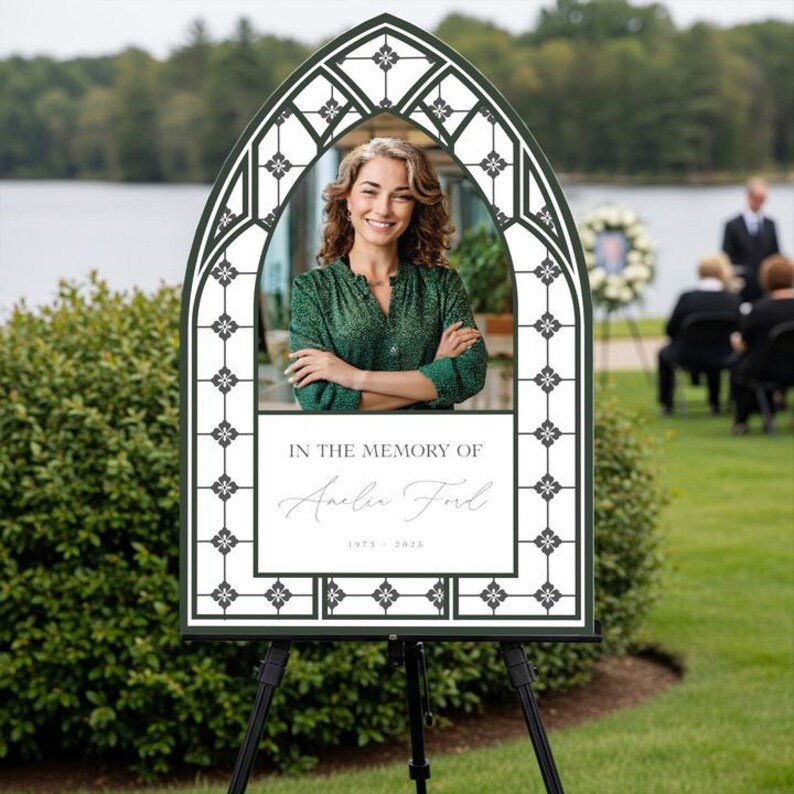 May include: Memorial sign with a photo of a woman in a green top, framed by a dark green and white decorative border. The sign reads "IN THE MEMORY OF" followed by a name and dates. The sign is displayed on a black easel outdoors.