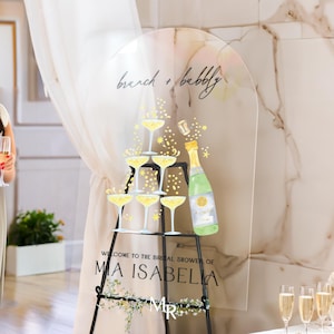 May include: Clear acrylic sign with a champagne tower and bottle illustration. The text reads "brunch + bubbly" and "Welcome to the bridal shower of Mia Isabella".